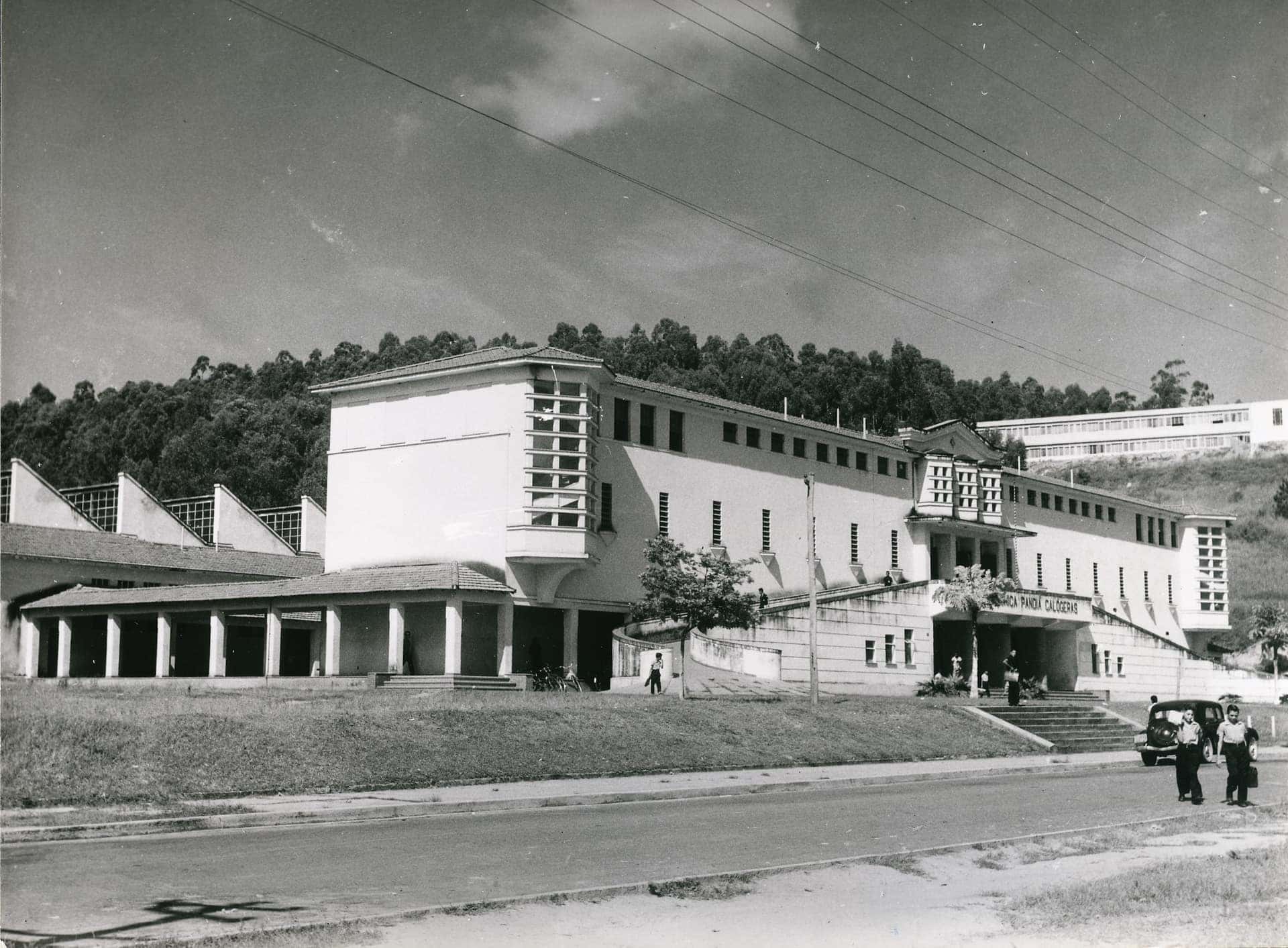 Fotografia de arquivo em preto e branco de um edifício escolar modernista com cobertura em shed e pessoas na calçada