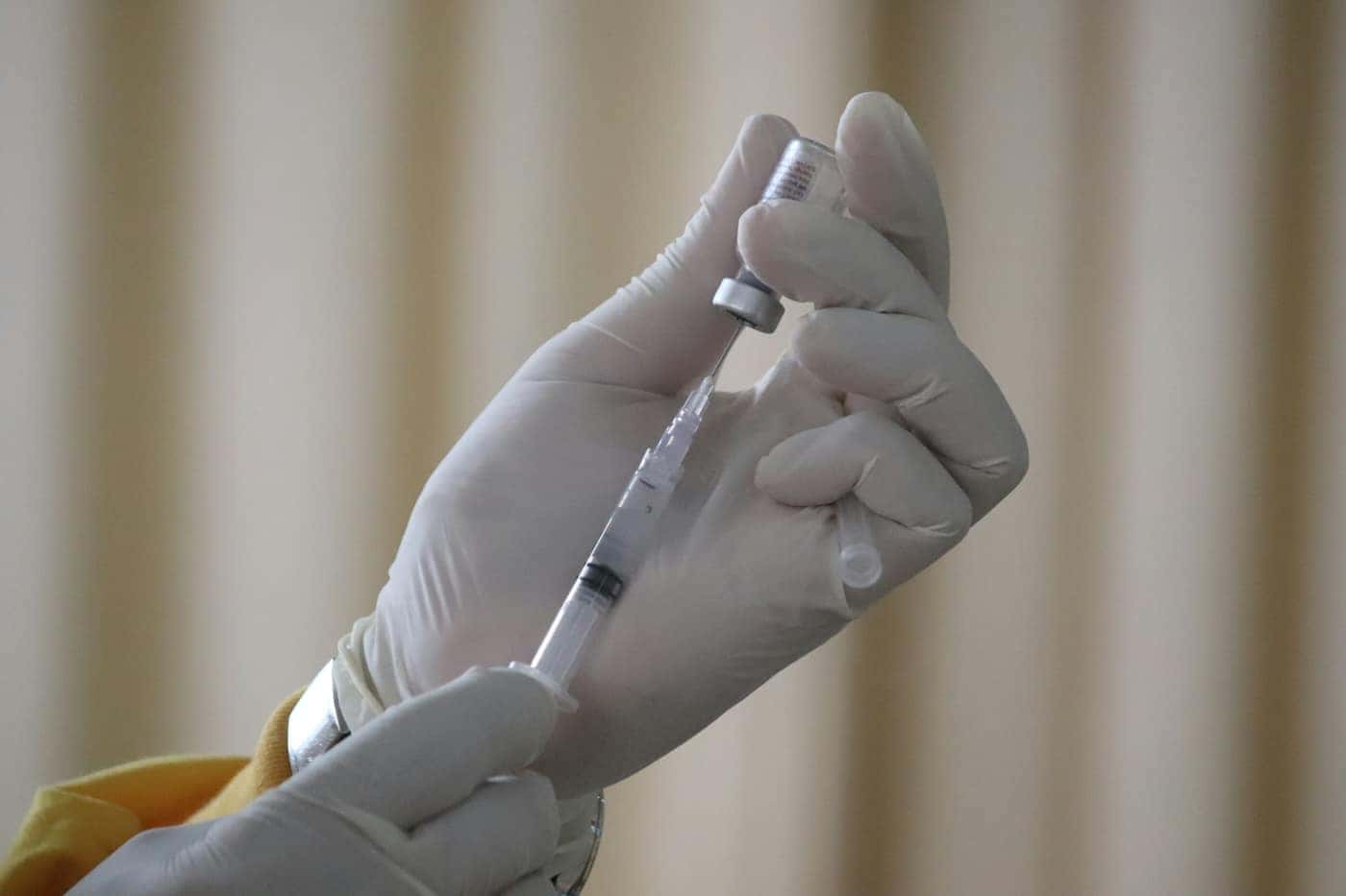 Mãos de luvas brancas retiram vacina de um frasco com uma seringa
